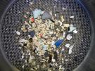 <p><strong>Fig. 5.24.</strong>&nbsp;Beach sand sifted in sieves to show (<strong>A</strong>) very coarse (2 mm diameter)&nbsp;sized sand grains.&nbsp;The blue, black, light green, and orange pieces are fragments of plastic debris.</p>
